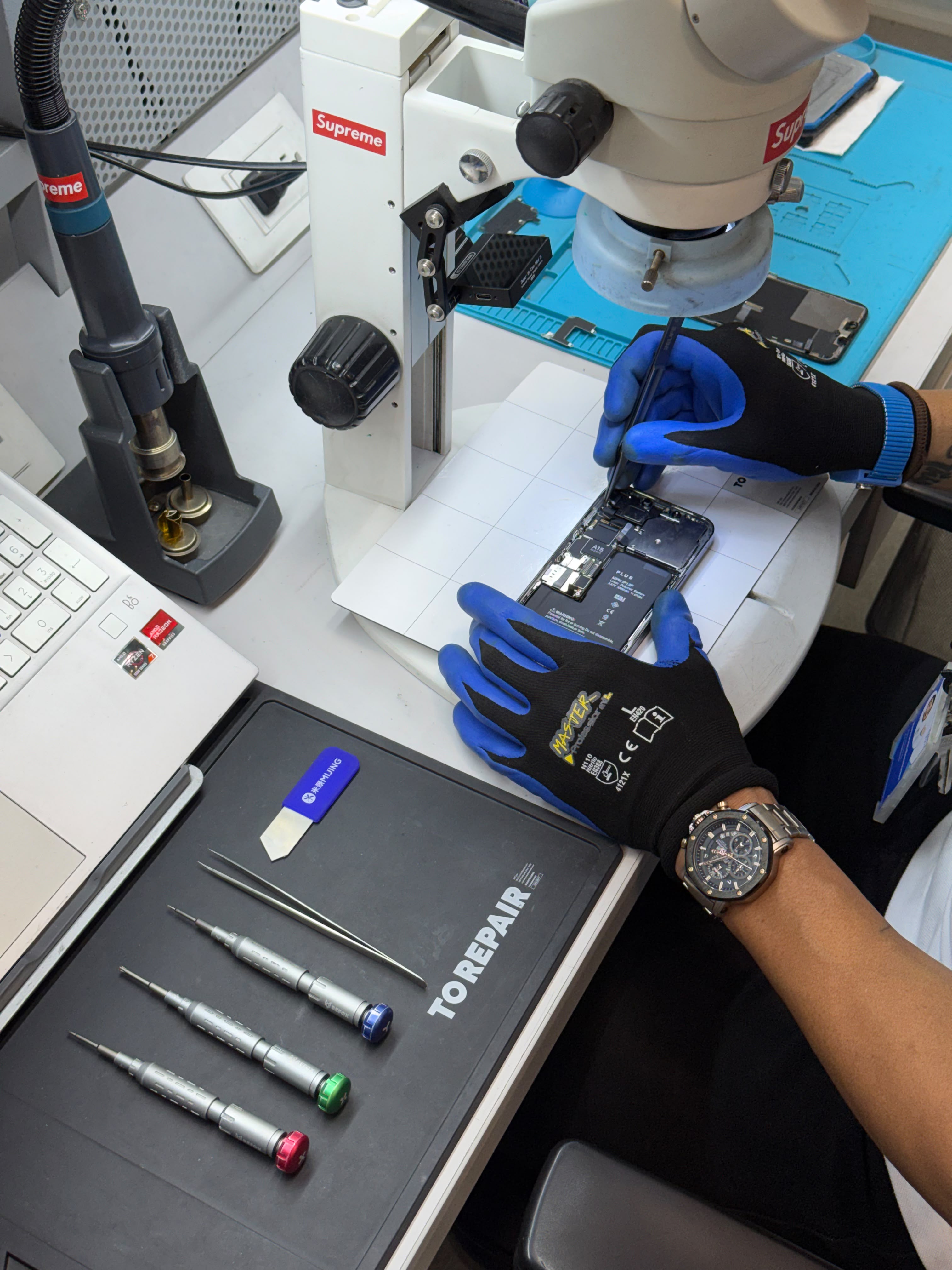 Manos con guantes de precisión reparando la placa lógica de un iPhone bajo el microscopio SmartFix